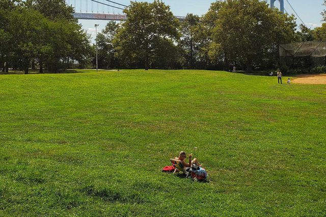 A man sits in a lawn chair on the green lawn of a Bay Ridge park near the Hudson River, with a view of the Verrazzano Narrows Bridge in the distance.